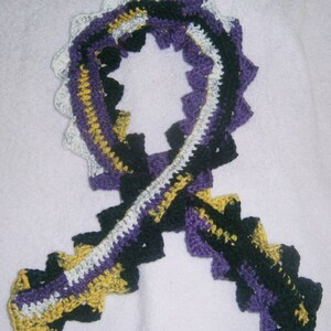 May include: A crocheted scarf in black, white, purple, and gold. The scarf is made in a repeating pattern of triangles and is tied in a loop.