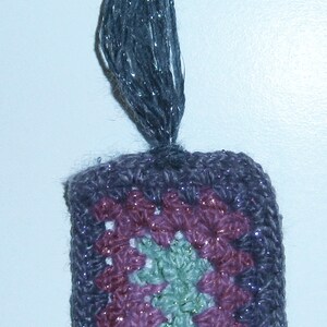 May include: A small, crocheted ornament with a purple, pink, and green pattern. The ornament has a long, gray tassel.