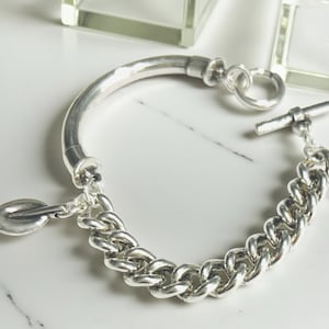 May include: A silver bracelet with a curved bar and a chain link design. The bracelet features a toggle clasp and a leaf-shaped charm. The jewelry is displayed on a white marble surface.