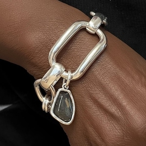 May include: A silver chain bracelet featuring large, polished rectangular links and a single, irregular, faceted dark gray stone pendant suspended from one of the links. The metal has a bright, reflective finish and a substantial, chunky appearance.