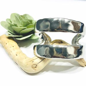 May include: Two silver-colored, hammered-look metal bracelets. The bracelets are stacked on top of each other. The bracelets are resting on a piece of driftwood, with a green succulent in the background. The background is white.