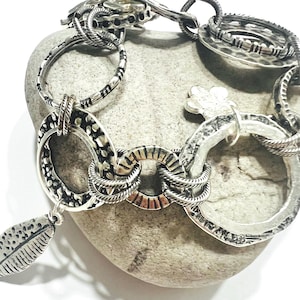 May include: A silver-toned bracelet with a variety of textured links, including oval and circular shapes. A leaf-shaped charm hangs from one link. The bracelet is displayed on a gray stone.
