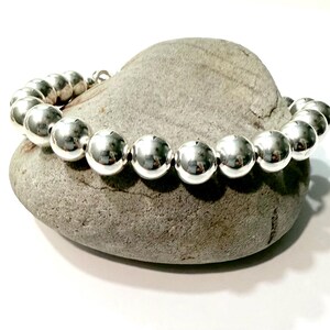 May include: A silver beaded bracelet with large, round beads. The bracelet is resting on a gray stone against a white background. The beads have a shiny, reflective surface.