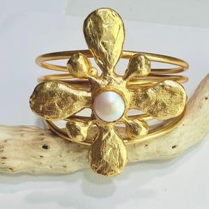 May include: A gold-toned cuff bracelet featuring a multi-layered wire band design. The centerpiece is a textured, flower-shaped ornament with five irregular petals and a central, smooth, round white pearl accent, creating a layered metallic look.