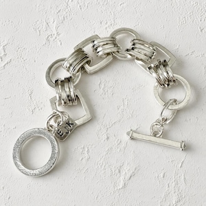 May include: A silver-toned chain bracelet featuring alternating circular and square links with a toggle clasp. A small rectangular charm attached to the bracelet is engraved with the letters EJK in a clear, sans-serif font.