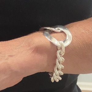 May include: A silver bracelet featuring a large, curved loop design connected to a chain of interwoven links. The bracelet is worn on a wrist, with the silver metal contrasting against a black garment.