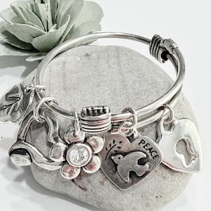 May include: A silver-toned expandable bangle bracelet featuring multiple charms including a leaf, a flower with a central clear crystal, a heart-shaped charm embossed with a bird and the word PEACE, and a heart-shaped charm with a keyhole cutout.