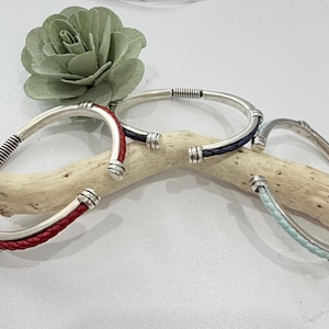 May include: Silver-toned metal bracelets with braided leather accents in red, navy blue, and light blue. The bracelets have a curved design with decorative metal ends and are displayed on a piece of driftwood.