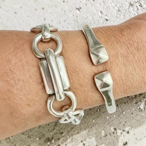 May include: A silver-toned link bracelet featuring elongated rectangular bars connected by circular rings, paired with a silver-toned cuff bracelet that has a smooth, polished finish and ends with two raised, pyramid-shaped square accents.