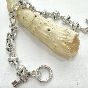 May include: A silver-toned bracelet with a key charm and a toggle clasp. The bracelet features a chain-link design with knotted details. The bracelet is resting on a piece of light-colored driftwood.