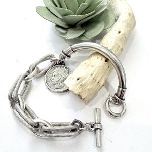 May include: A silver-toned bracelet with a toggle clasp and a curved bar. The bracelet features a chain-link design and a circular charm with a profile image. The bracelet is displayed with a piece of driftwood and a green decorative element.