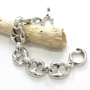 Peut inclure: Un bracelet argenté avec un design de chaîne nautique. Le bracelet est doté d'un fermoir à bascule et est présenté sur un morceau de bois flotté sur fond blanc. Les maillons mesurent environ 2 cm de long.