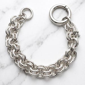 May include: A silver-toned chain bracelet featuring a dense, interlocking circular link pattern. The bracelet is secured with a large, round spring-ring clasp at one end and a smaller circular jump ring at the other, creating a continuous, heavy-duty look.