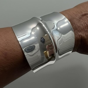 May include: A wide, polished silver cuff bracelet with a curved design. The bracelet is worn on a wrist and reflects the surrounding environment. The metal has a bright, shiny appearance.