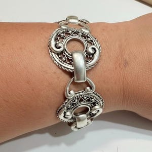 May include: A silver-toned bracelet featuring repeating circular links with intricate cut-out heart patterns and swirling scrollwork details. The design includes smooth, solid rectangular connectors between each ornate circular element.