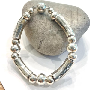 May include: A silver bracelet featuring a mix of round and cylindrical beads. The bracelet has a magnetic clasp and is displayed on a gray stone. The beads have a polished, reflective surface, giving the bracelet a modern look.