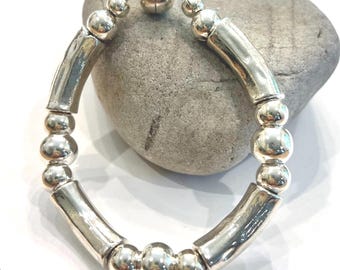 May include: A silver bracelet featuring a mix of round and cylindrical beads. The bracelet has a magnetic clasp and is displayed on a gray stone. The beads have a polished, reflective surface, giving the bracelet a modern look.