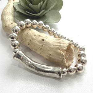 May include: Silver beaded bracelet with a cylindrical bar accent. The bracelet features large, round beads and a smooth, polished finish. The jewelry is displayed on a piece of driftwood and a green paper flower.