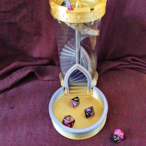 May include: A gold and silver tower-shaped dice rolling tower with a clear plastic tube and a spiral staircase inside. Four multi-colored dice with black and gold numbers are scattered around the tower.