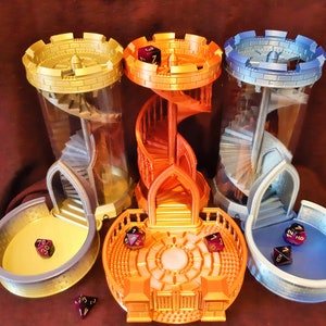 May include: Three 3D printed dice towers, each with a different color scheme: gold, orange, and blue. The towers are designed to look like miniature castles with spiral staircases inside. Each tower has a matching dice tray at its base.