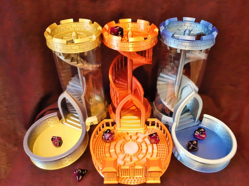 May include: Three 3D printed dice towers, each with a different color scheme: gold, orange, and blue. The towers are designed to look like miniature castles with spiral staircases inside. Each tower has a matching dice tray at its base.
