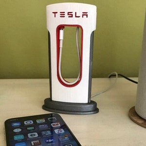 Może przedstawiać: Biały stojak do stacji ładowania Tesla, wydrukowany w 3D, z czerwonym akcentem i napisem "TESLA" nadrukowanym na czerwono. Biały kabel jest przewleczony przez stojak. Smartfon leży na stole przed stojakiem.