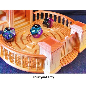 May include: A gold-colored 3D printed tray with a circular courtyard design. The tray has a gate and a balcony with railings. There are three iridescent dice on the tray, one blue, one purple, and one green.