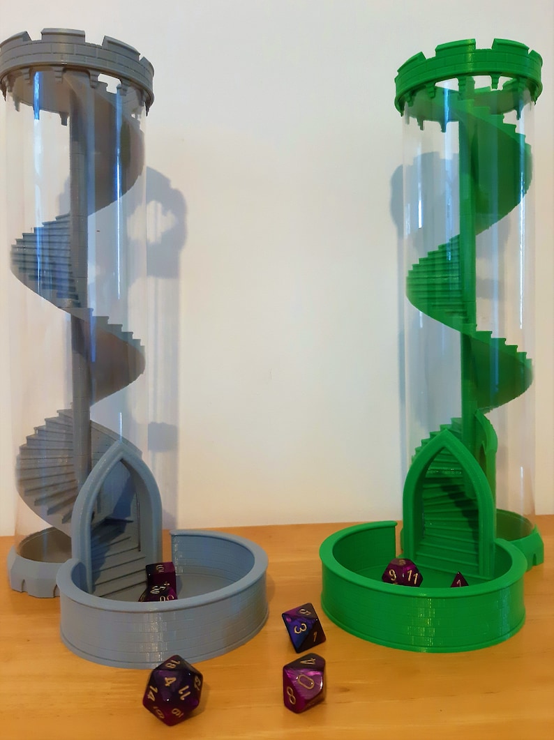 May include: Two 3D printed dice towers, one gray and one green, with clear acrylic tubes and spiral staircases. The towers are on a wooden surface with purple dice scattered around them.