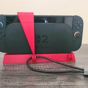 以下が含まれることがあります： 赤色のスタンドに固定された黒色のNintendo Switchコンソール。ロックケーブルとキーが付属しています。コンソールには青とオレンジのボタンがあります。スタンドには「D2 Nintendo Switch」の文字があります。