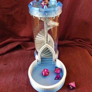 May include: A blue and silver 3D printed dice tower with a spiral staircase inside. The tower has a removable tray at the bottom to catch the dice. Four purple and gold dice are scattered around the tower.