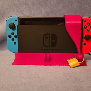 Può includere: Una console Nintendo Switch nera e rosa è assicurata con un lucchetto dorato e un supporto rosa. La console è in modalità da tavolo con lo schermo rivolto in avanti. La console è collegata a un cavo nero.