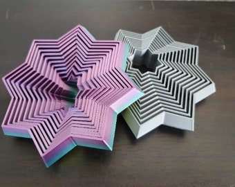 Puede incluir: Dos objetos decorativos en forma de estrella. Uno es un degradado de morado, azul y verde. El otro es gris. Ambos tienen un diseño en zigzag en capas. Los objetos están sobre una superficie marrón oscuro.