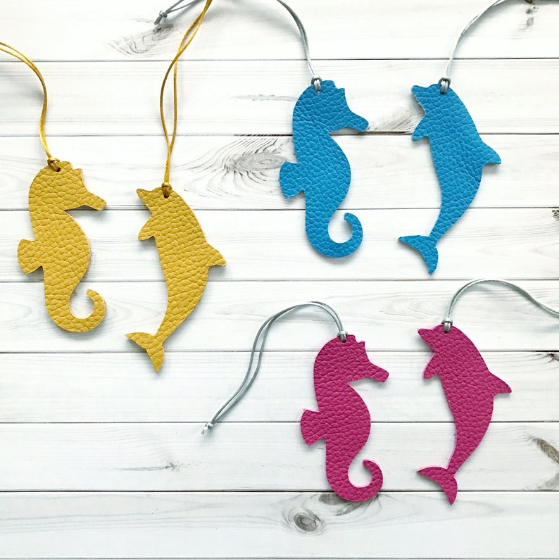 May include: Six leather seahorse and dolphin ornaments in blue, yellow, and pink. The ornaments are hanging from strings on a white wooden background.