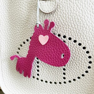 May include: A pink leather giraffe charm with a pink heart on its chest. The charm is attached to a white leather bag with a perforated design.