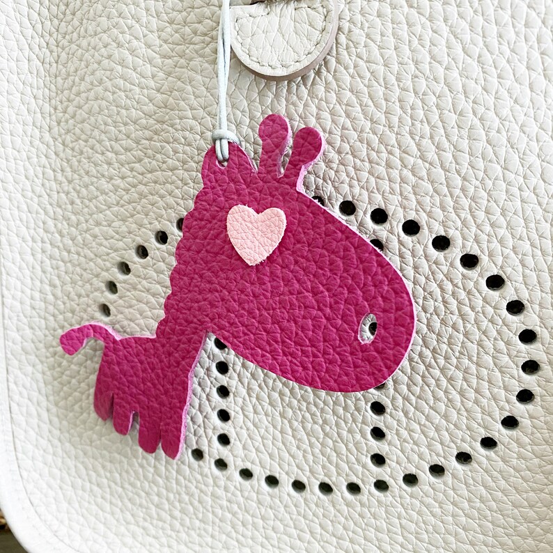 May include: A pink leather giraffe charm with a pink heart on its chest. The charm is attached to a white leather bag with a perforated design.