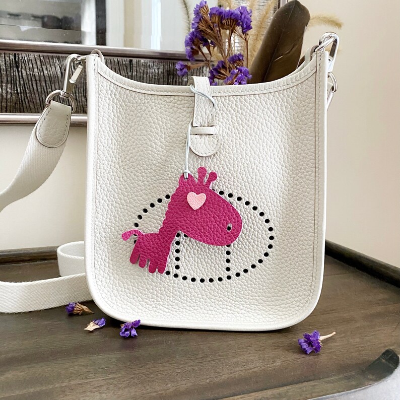 May include: A white leather shoulder bag with a pink giraffe charm. The bag has a perforated design and a white strap.