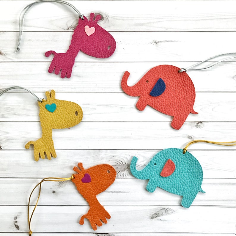 May include: Four leather animal ornaments with heart-shaped eyes. The ornaments are a pink giraffe, a yellow giraffe, a red elephant, and a blue elephant. Each ornament has a gold string attached.