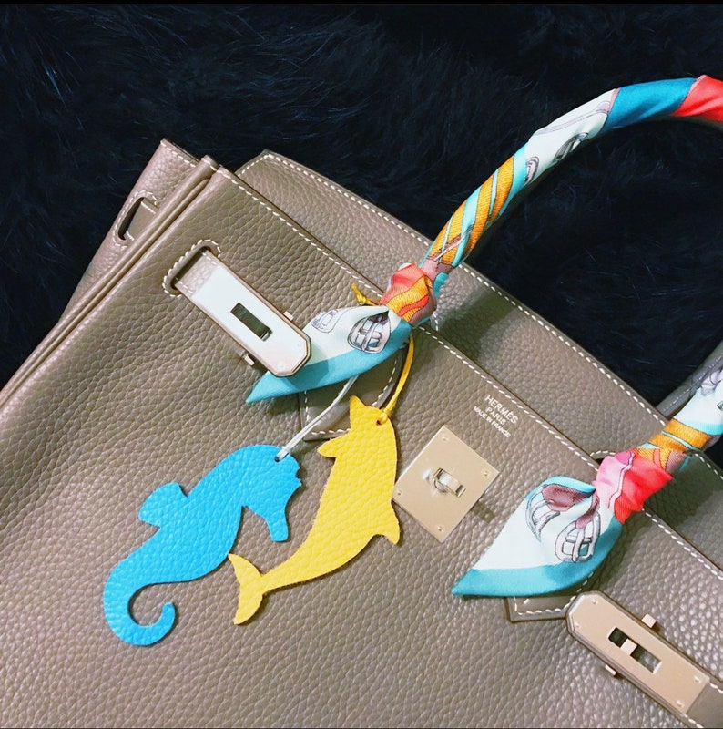 May include: A brown leather handbag with a gold clasp and a colorful scarf with a seahorse and dolphin charm attached. The handbag has the text "HERMES PARIS MADE IN FRANCE" on the front.