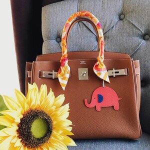 May include: A brown leather Birkin style handbag with gold hardware and a colorful patterned scarf tied around the handles. The handbag has a small red elephant charm attached to it.
