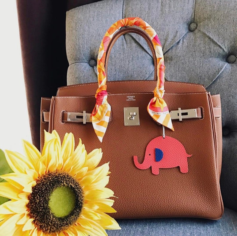 May include: A brown leather Birkin style handbag with gold hardware and a colorful patterned scarf tied around the handles. The handbag has a small red elephant charm attached to it.