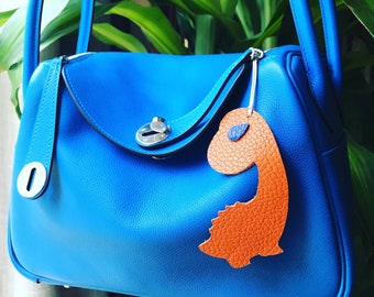 May include: A bright blue leather handbag with a top flap closure and a silver clasp. The bag has a small orange leather dinosaur charm attached to it.