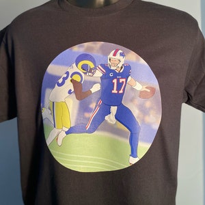 May include: Black t-shirt with a circular graphic of two football players in action. The player on the left is wearing a blue and yellow uniform with the number 30. The player on the right is wearing a blue uniform with the number 17.