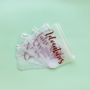May include: Six clear plastic zippered pouches with holes for a binder. The pouches have the words "Adventures", "Take", "Dream", "Create", "Love", and "Explore" printed in maroon.