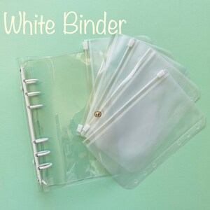 Op de afbeelding: Een doorzichtige plastic map met een ritsvak. De map heeft zes ringen en het vak heeft een witte rits. De tekst "White Binder" staat bovenaan de afbeelding in het geel.