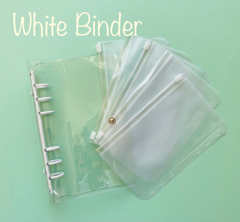 Op de afbeelding: Een doorzichtige plastic map met een ritsvak. De map heeft zes ringen en het vak heeft een witte rits. De tekst "White Binder" staat bovenaan de afbeelding in het geel.
