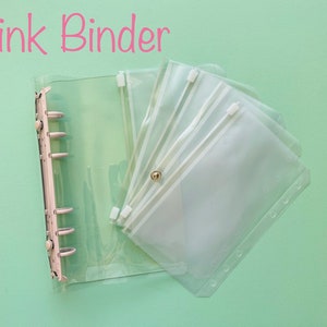 Op de afbeelding: Een doorzichtige plastic map met een ritsvak en meerdere doorzichtige plastic vakken. De map heeft een ringmechanisme en de tekst "Pink Binder" staat in roze letters bovenaan de afbeelding.