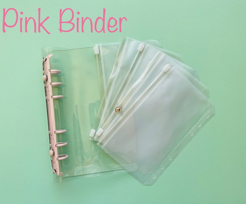 Op de afbeelding: Een doorzichtige plastic map met een ritsvak en meerdere doorzichtige plastic vakken. De map heeft een ringmechanisme en de tekst "Pink Binder" staat in roze letters bovenaan de afbeelding.