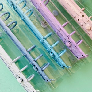Op de afbeelding: Vier doorzichtige plastic ringbanden met verschillende gekleurde ringen. De ringen zijn lichtblauw, blauw, paars en wit. De ringbanden zijn op elkaar gestapeld.