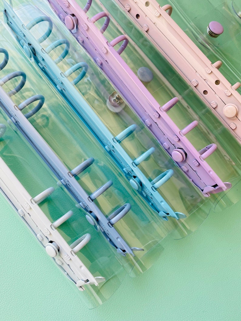 Op de afbeelding: Vier doorzichtige plastic ringbanden met verschillende gekleurde ringen. De ringen zijn lichtblauw, blauw, paars en wit. De ringbanden zijn op elkaar gestapeld.