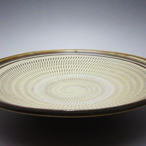 May include: A ceramic plate with a cream-colored center and a brown rim. The plate features a radial pattern of thin, dark lines emanating from the center. The rim has a dark brown outer edge and a lighter brown inner band.